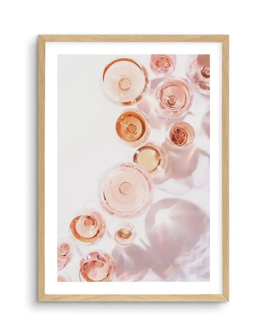 Framed photography art print featuring an overhead view of multiple champagne glasses filled with rosé champagne, displayed in a natural oak frame with white border. The glasses are arranged organically on a white surface, casting soft pink shadows and reflections, creating a light and celebratory mood.