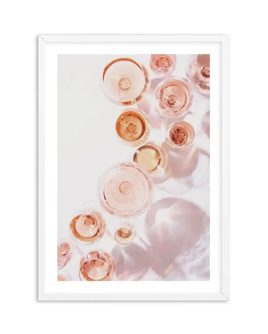 Framed photography art print of multiple champagne glasses filled with rosé champagne, viewed from directly above, on a white background with soft pink shadows. The composition features various sizes of round glasses with reflections and refractions of light, displayed in a white frame with white border.