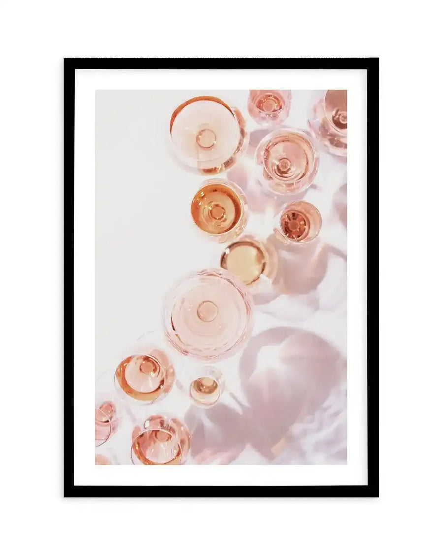 Framed art print featuring an overhead photographic composition of multiple champagne glasses filled with rosé champagne, casting soft pink shadows on a white surface, with a black frame and white border.