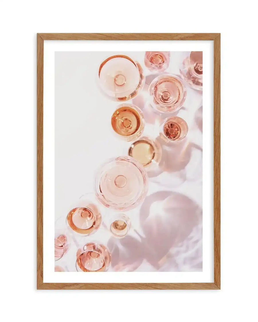 "Framed art print featuring an overhead photograph of multiple champagne glasses filled with rosé champagne, displayed in a natural oak frame with white border. The glasses are arranged in a scattered composition on a white surface, casting soft pink shadows and reflections."