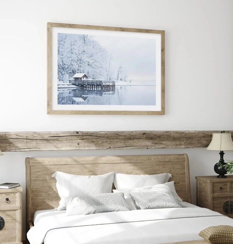 Lifestyle shot of a bedroom featuring a large framed art print above a wooden headboard. The print is a photograph of a serene winter lake scene with a small wooden boathouse on a pier, surrounded by snow-covered trees and a calm, reflective body of water under a soft, overcast sky. The print has a natural wood frame and a white border. The bedroom has a rustic aesthetic with a wooden headboard, white bedding, and wooden bedside tables with lamps.