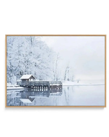 Framed canvas print of a serene winter landscape featuring a small wooden cabin and pier on the edge of a frozen lake, surrounded by snow-covered trees, with a natural oak frame. The scene is rendered in cool tones of white, grey, and pale blue, with reflections of the cabin and trees visible in the still water.
