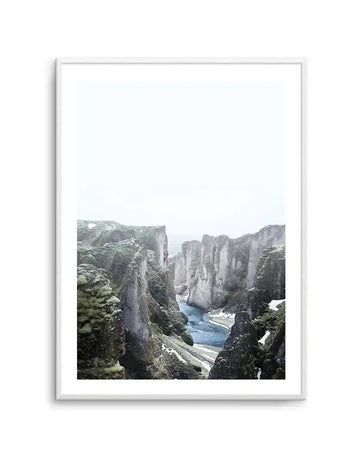 Framed art print of a misty mountain landscape featuring towering grey stone cliffs with moss-green vegetation, a narrow blue stream flowing through the rocky gorge below, and patches of snow on surrounding peaks. The photograph has a soft, overcast quality with muted colors dominated by grey, sage green, and pale blue tones. The print is mounted with a white border in a light grey frame.