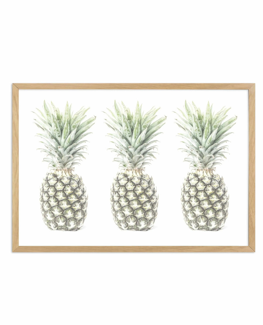 Three pineapples rendered in detailed watercolor botanical illustration, arranged horizontally across a white background. Each pineapple features a textured, pale green and cream-colored body with characteristic diamond-patterned segments, topped with a crown of long, pointed green leaves. The artwork is displayed in a natural wood frame with a white border, creating a clean, minimalist presentation against a neutral background.