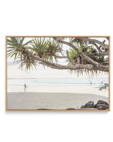 Framed canvas art print featuring a coastal beach scene with a surfer walking along the wet sand. The composition captures a wide expanse of light grey sand, gentle white waves, and a pale sky. In the foreground, a large tree with thick, gnarled branches and spiky green leaves extends across the top of the frame. The print is displayed in a natural oak frame.