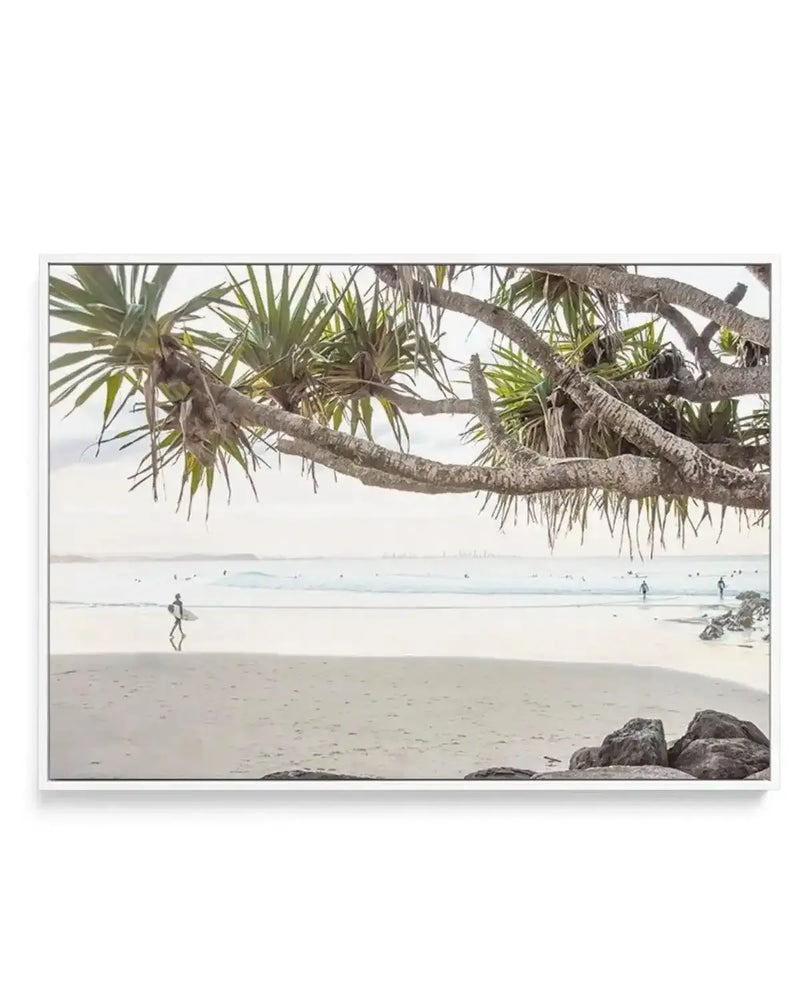 Framed canvas print of a coastal beach scene with a large, gnarled tree branch with long, spiky green leaves extending across the top of the frame. Below the branch, a lone surfer walks along wet sand at low tide, carrying a surfboard, with gentle waves and an overcast sky stretching to the horizon. Several other surfers are visible in the distance in the water, and rocks are visible in the foreground on the right. The print is displayed in a white frame.