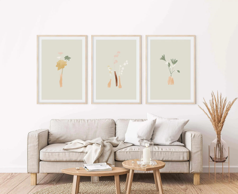Three framed art prints displayed above a light beige sofa in a minimalist living room. Each print features a different abstract floral design in soft, muted tones of green, yellow, pink, and brown on a pale sage green background, with natural wood frames and white borders. The room includes a woven rug, a wooden coffee table with a candle, and dried wheat stalks in a glass vase.