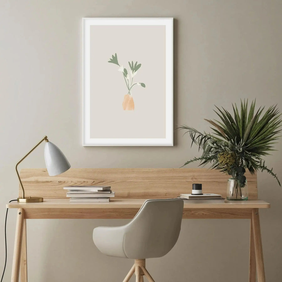 Lifestyle shot of a minimalist home office with a Le Posy III art print displayed above a light wood desk. The print features a minimalist illustration of three white flowers with sage green leaves in a small, abstract peach-colored vase, set against a warm beige background with a white frame and white border. The desk has a white lamp with a gold base, a stack of books, and a glass vase with various green plants. A light grey office chair is pulled up to the desk.