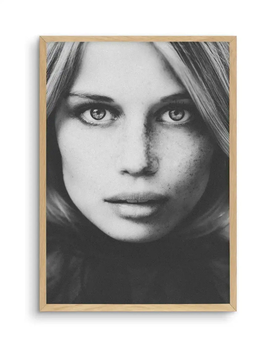 Framed black and white photograph of a woman's face in a natural oak frame with white border. The close-up portrait features a woman with long, straight blonde hair framing her face, looking directly at the viewer with a serious expression. Her eyes are light, and her face is lightly freckled, with a dark turtleneck visible below her chin.