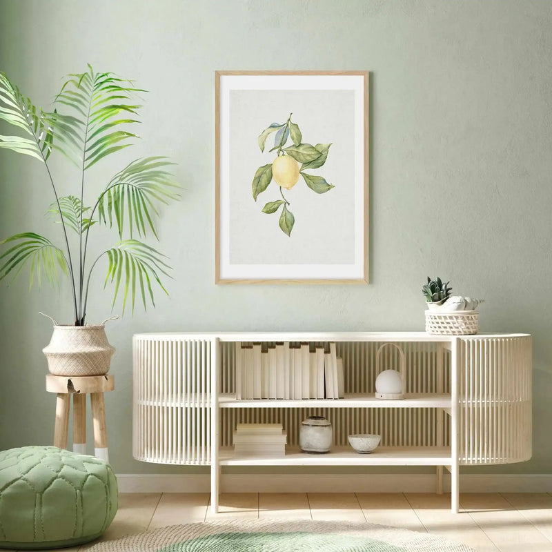 Lifestyle shot of an art print displayed in a minimalist living room with a light sage green wall. The framed art print features a watercolor illustration of a single lemon on a branch with green leaves, set against a light cream background, with a natural wood frame and white border. Below the print is a white slatted console table with books and decorative items. To the left, a large potted palm plant sits on a wooden stool, and a light green pouf rests on the floor.