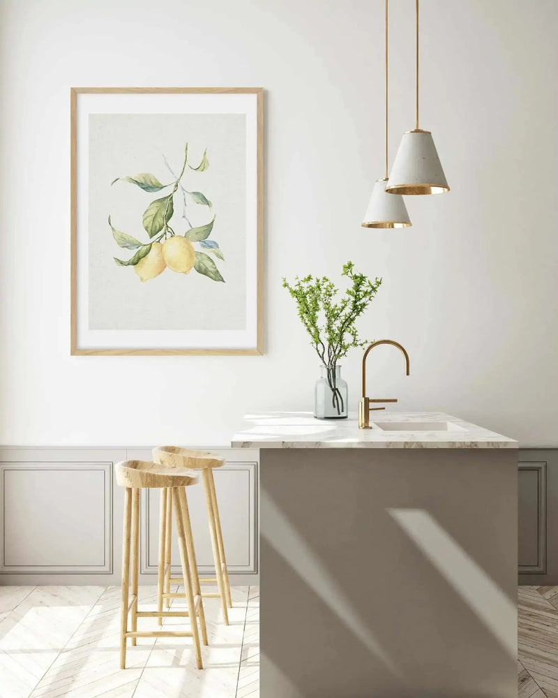 Kitchen:
Large framed art print displayed above a kitchen island with two wooden bar stools. The print features two lemons on a branch with green leaves against a cream background, with natural wood frame and white border. The kitchen has white walls with wainscoting, a gold faucet, and two pendant lights with concrete shades and gold accents.