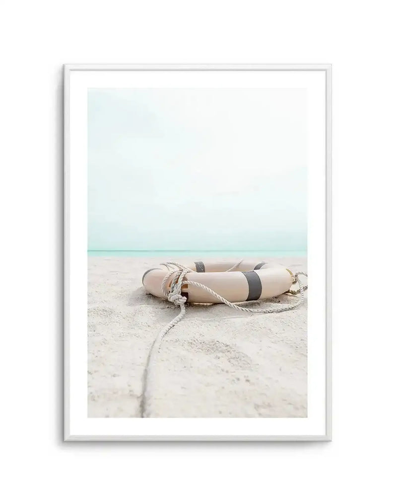 Framed photography art print of a lifebuoy resting on a sandy beach, with a white frame and white border. The lifebuoy is light beige with two dark grey stripes and a rope tied around it, extending into the foreground. In the background, a calm, light blue ocean meets a pale, almost white sky.
