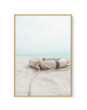 Framed canvas art print featuring a weathered cream and gray lifebuoy with coiled rope lying on pale sand beach, with turquoise ocean water and soft blue-gray sky in the background. The photograph has a calm, minimalist aesthetic with muted tones. Natural wood frame with white border.