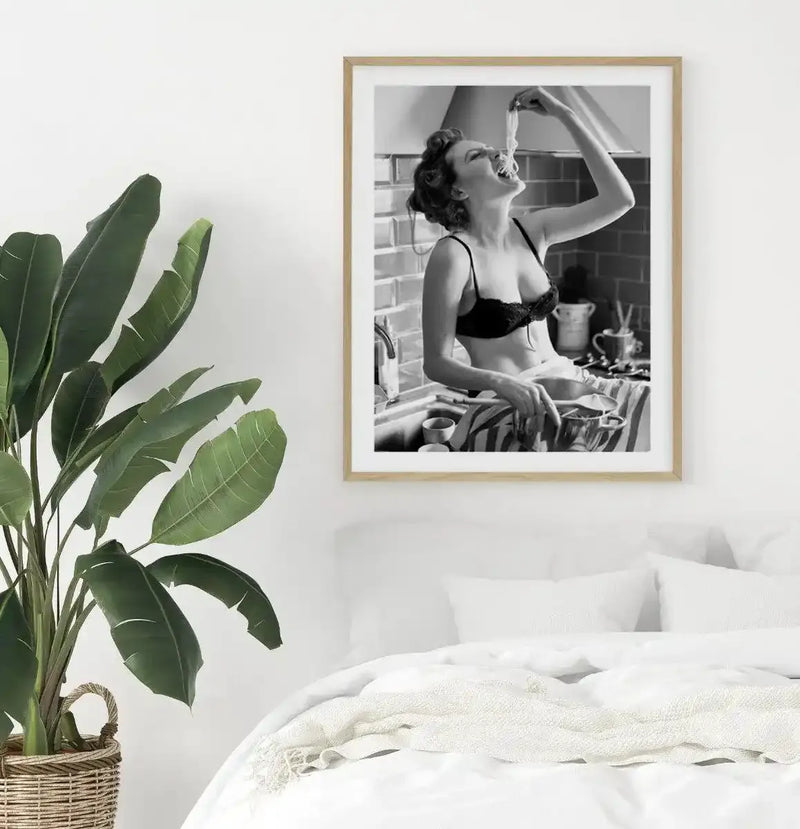 Lifestyle shot of a bedroom with a large framed black and white art print displayed above a white bed. The print features a woman in a black bra sitting on a kitchen counter, joyfully eating spaghetti with her hands. The room includes a large green plant in a woven basket on the left and a white bed with a textured cream throw.