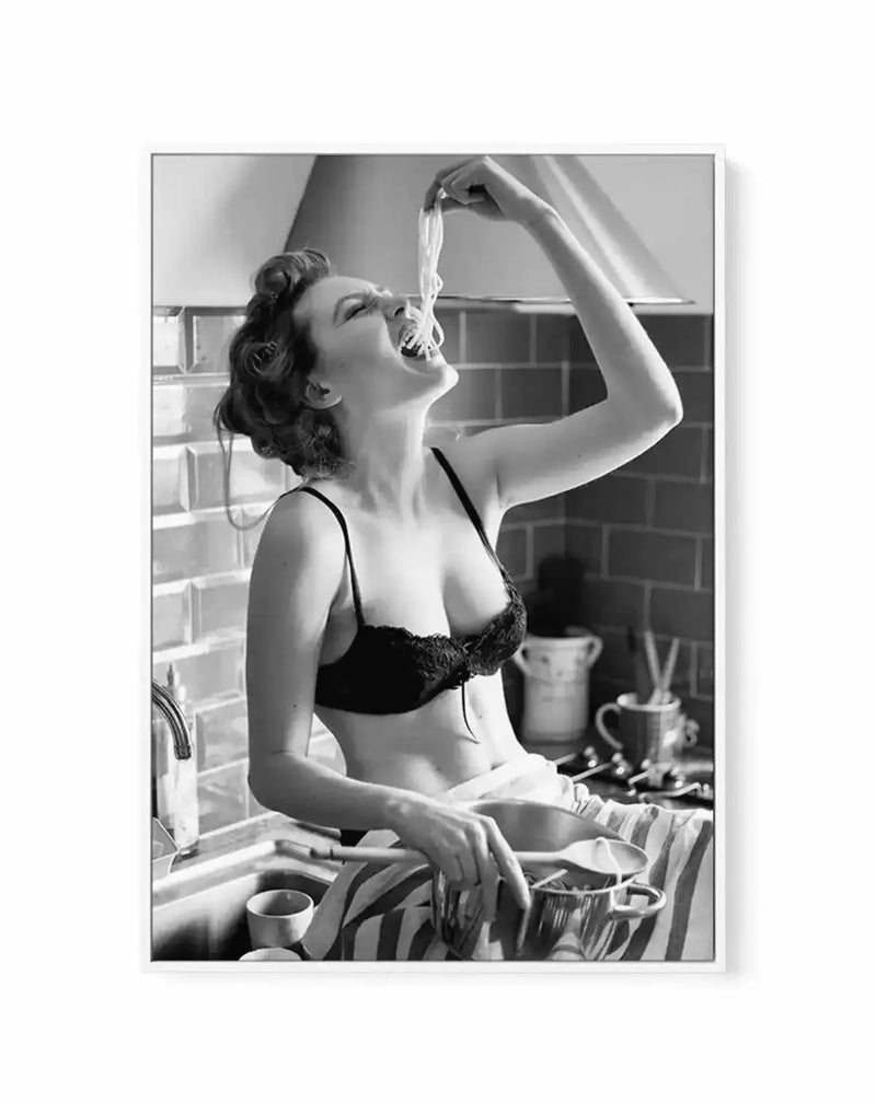 Black and white vintage photograph framed as canvas art print with white border. Image shows a woman in 1940s style at a kitchen sink, wearing black lingerie, holding a strand of pasta up to her mouth with a joyful expression. Kitchen setting visible with dishes, cups, and window in soft-focus background. Glossy black and white photography with classic mid-century aesthetic. Frame appears to be natural wood or light finish. Canvas print mounted on white-bordered frame.