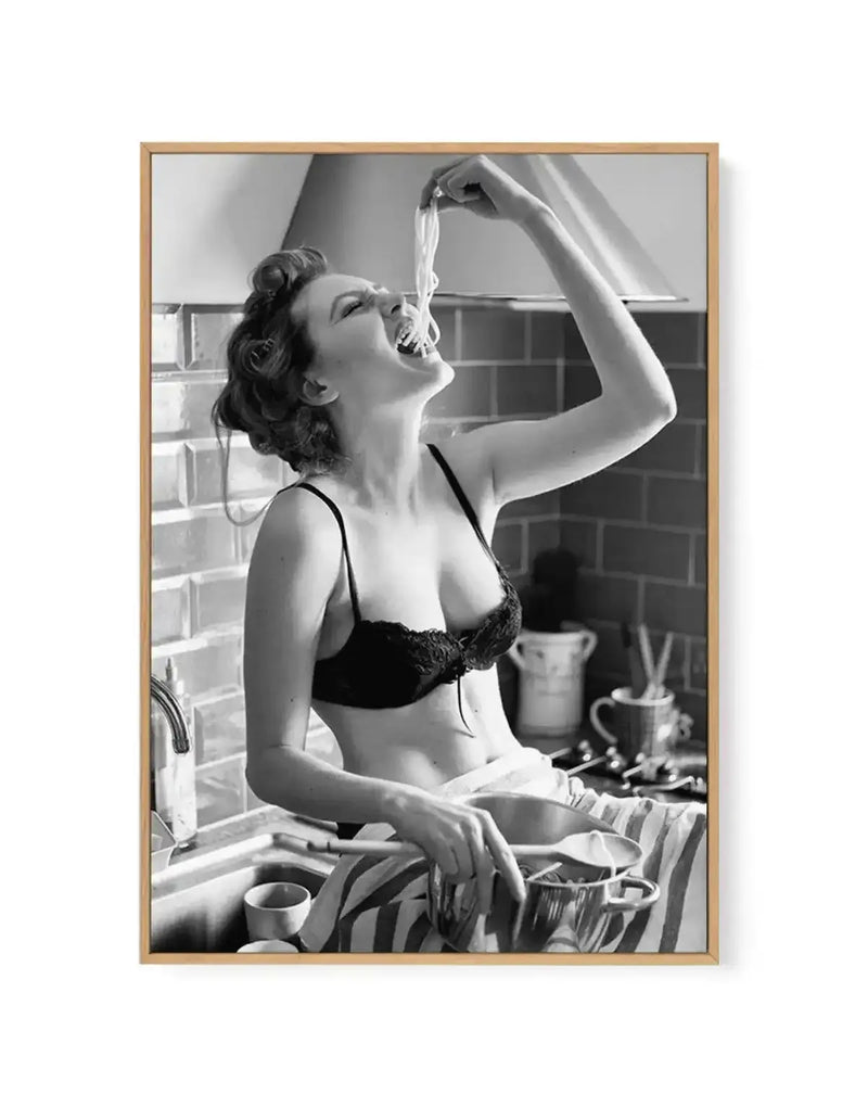 Black and white vintage photograph framed in natural wood showing a woman at a kitchen sink wearing dark lingerie, tilting her head back with a piece of fabric in her hand, surrounded by dishes and kitchenware, embodying a playful, carefree mid-century aesthetic. Canvas print with white border.