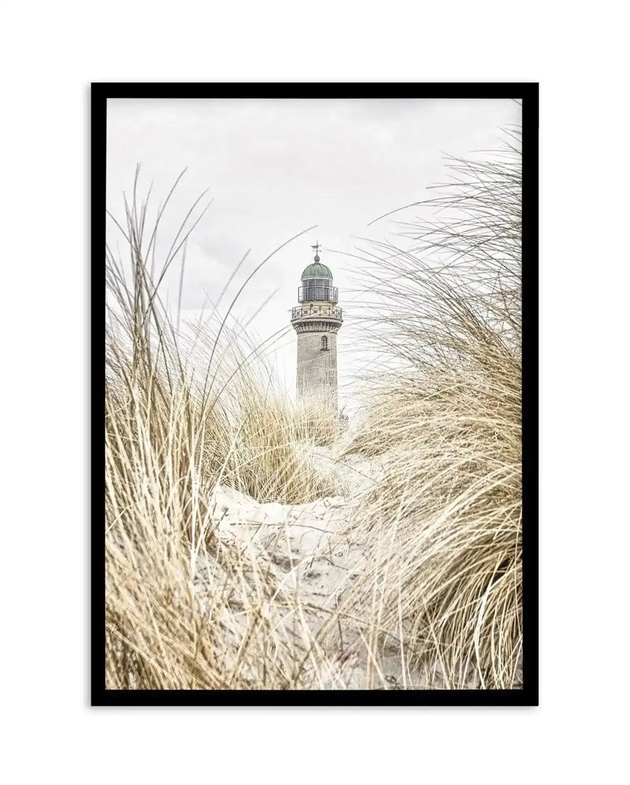 Framed black and white photograph of a coastal lighthouse viewed through tall, dry dune grass, with a black frame and white border. The composition features a stone lighthouse with a dark green dome, partially obscured by the golden-brown grass and sand dunes in the foreground, under an overcast sky.