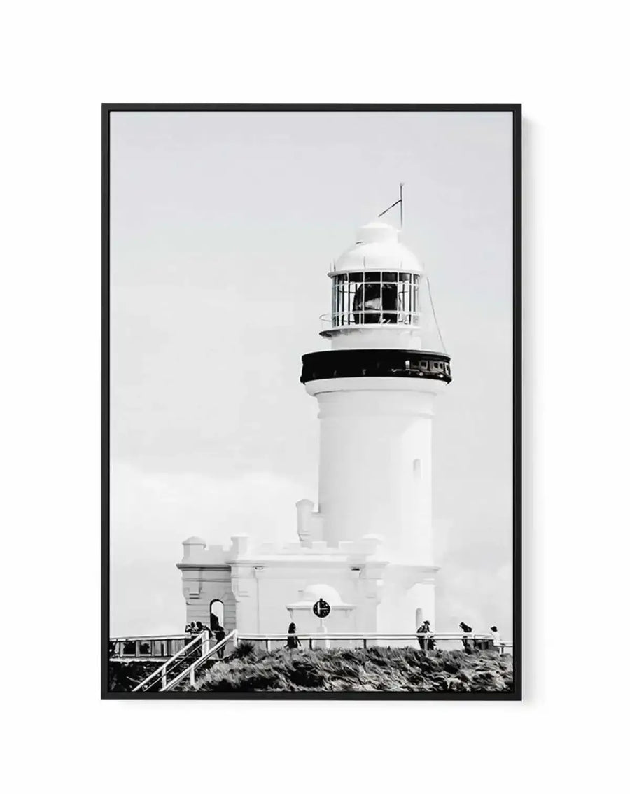 Framed black and white photograph of the Byron Bay Lighthouse with a black frame and white border. The composition features the white lighthouse against a bright, overcast sky, with people visible on a grassy hill and wooden walkway in the foreground.
