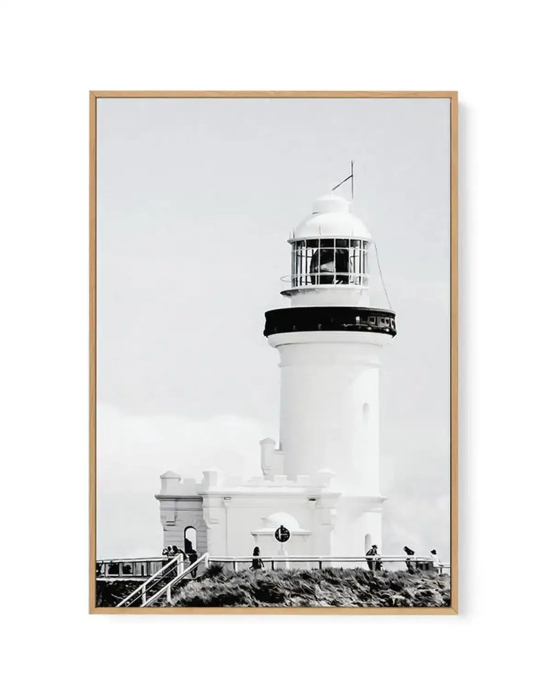 Black and white framed canvas print of Byron Bay Lighthouse. The photograph shows a tall white cylindrical lighthouse tower with a black band around its middle section and an enclosed lantern room at the top, topped with a flagpole. The base features white keeper's quarters with a railing and wooden boardwalk. Several visitors stand on the viewing platform. Sparse coastal vegetation and overcast sky create a minimalist composition. Natural wood frame with white border.