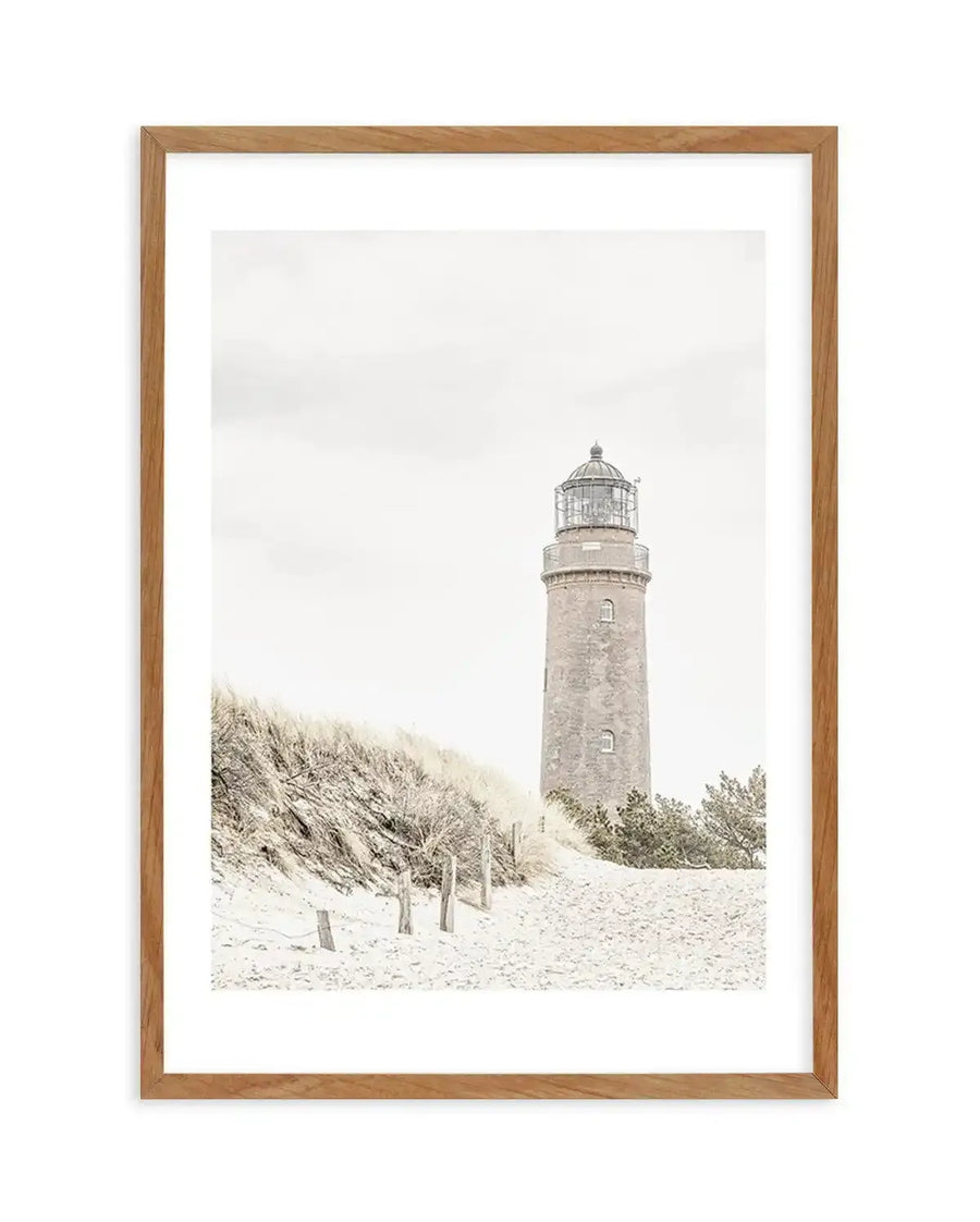 Framed black and white photograph of a lighthouse on a sandy beach with natural wood frame and white border. The composition features a tall, slender lighthouse with a domed top, standing behind a dune covered in dry grass and sparse vegetation. Wooden posts are visible in the sand in the foreground.