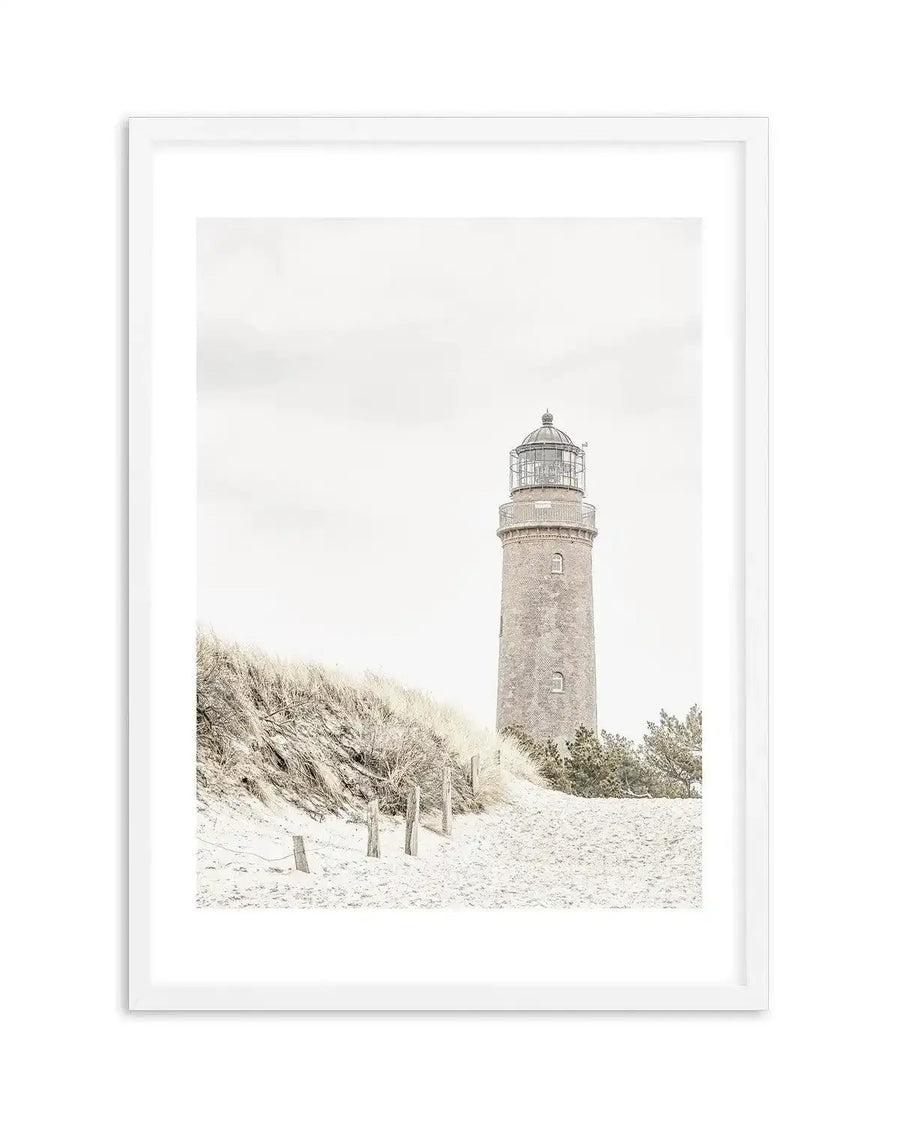 Framed photography art print of a coastal lighthouse on a sandy dune, with a white frame and white border. The composition features a tall, brick lighthouse with a dark metal lantern room, standing behind a sandy dune covered in dry, light-colored grasses and sparse shrubs. A wooden fence with posts and rope runs along the base of the dune. The sky is a bright, overcast white, creating a soft, muted light over the scene.