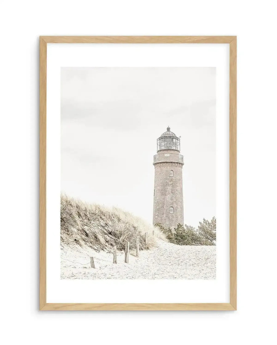 Framed black and white photograph of a coastal scene with a lighthouse and sand dunes, displayed in a natural oak frame with white border. The composition features a tall, brick lighthouse on the right, with sandy dunes covered in dry grass and wooden posts on the left, under a bright, overcast sky.