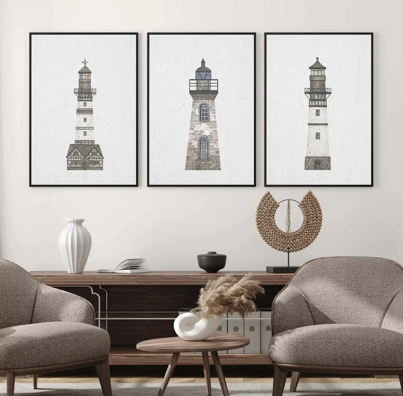 Framed canvas art print featuring a detailed architectural illustration of a lighthouse with a stone base, white tower, and lantern room at the top. The drawing uses gray and tan tones rendered in a fine-line illustration style. The piece is displayed in a black frame with white border, hung on a cream wall above a mid-century modern living room setting with two upholstered gray chairs, a wooden side table, and decorative objects including a white ceramic vase and woven wall sculpture.