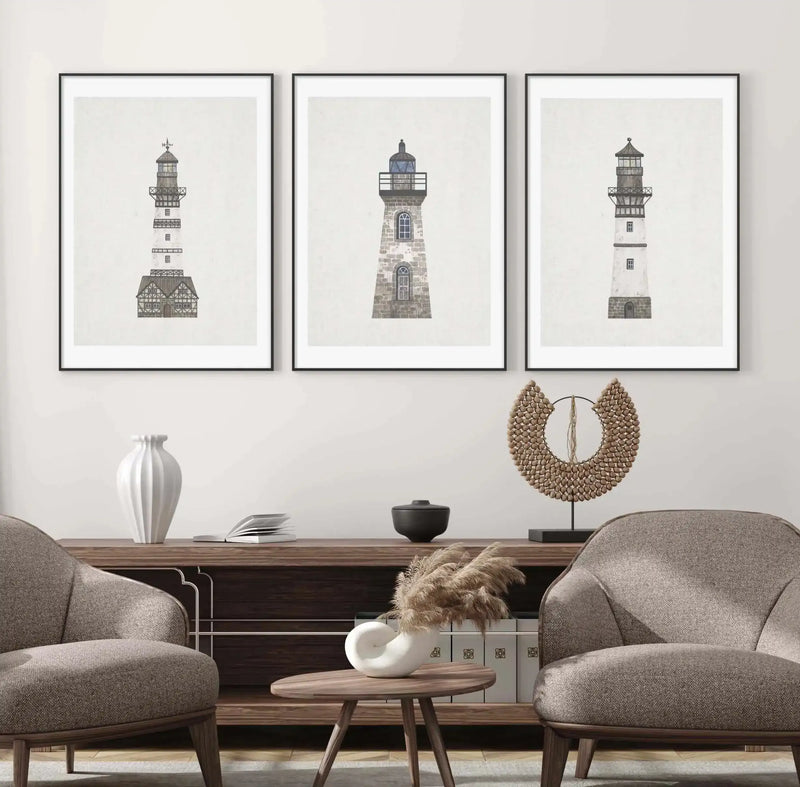 Lifestyle shot of three framed art prints displayed above a dark wood console table in a neutral-toned living room. Each print features a different lighthouse illustration in muted grey tones on a light cream background, with black frames and white borders. The room includes two upholstered armchairs, a white fluted vase, a black lidded bowl, a decorative beaded sculpture, and a small round wooden side table with a white spiral vase holding dried pampas grass.