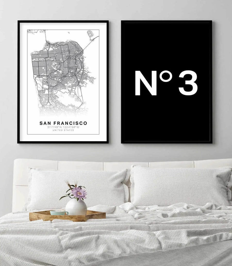 Lifestyle shot of a bedroom featuring two framed art prints above a white bed. The left print is a line art map of San Francisco in black on a white background, with a black frame and white border. The right print is a black background with the text "N°3" in white, with a black frame and white border. The bed has white pillows and a white textured blanket, with a wooden tray holding a vase with purple flowers and a light blue cup.