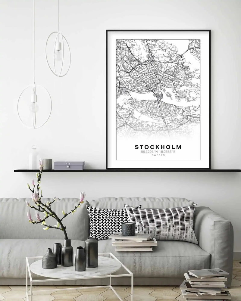 Lifestyle shot of a minimalist living room with a framed art print of a Stockholm map displayed above a grey sofa. The print features a black and white line art map of Stockholm with major roads and waterways, and the text "STOCKHOLM 59.3293°N, 18.0686°E SWEDEN" at the bottom, all in a black frame with a white border. The room includes a black floating shelf with decorative items, a white marble coffee table with dark grey vases, and black and white patterned cushions on the sofa.