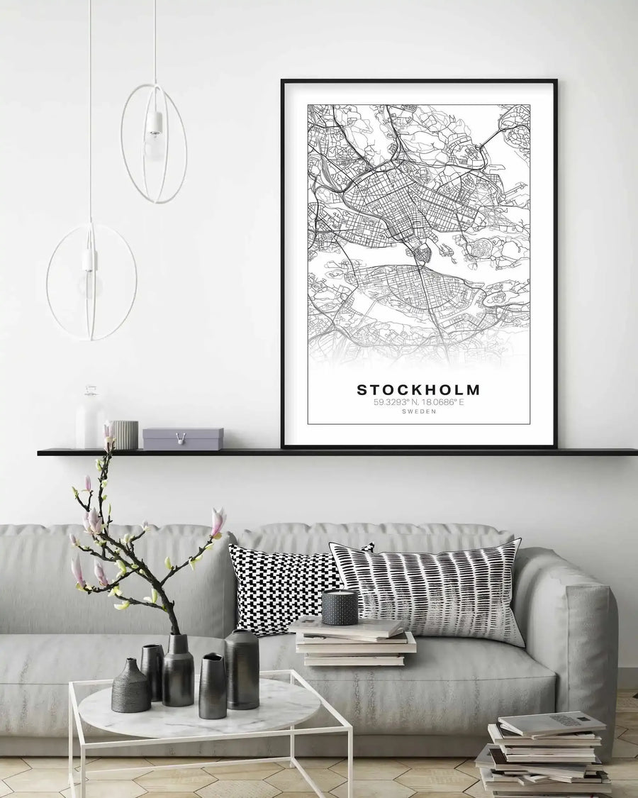 Lifestyle shot of a minimalist living room with a framed art print of a Stockholm map displayed above a grey sofa. The print features a black and white line art map of Stockholm with major roads and waterways, and the text "STOCKHOLM 59.3293°N, 18.0686°E SWEDEN" at the bottom, all in a black frame with a white border. The room includes a black floating shelf with decorative items, a white marble coffee table with dark grey vases, and black and white patterned cushions on the sofa.
