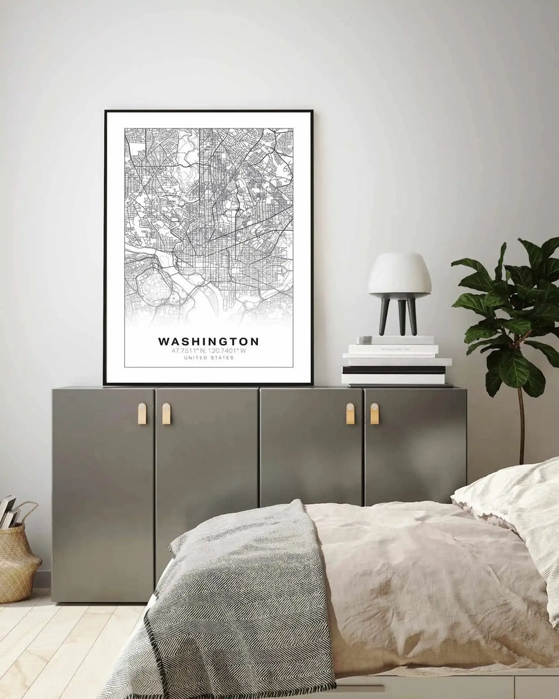 Lifestyle shot of a bedroom with a framed art print displayed above a grey cabinet. The print features a black and white line art map of Washington, with the city name and coordinates below, displayed in a black frame with a white border. The room includes a white lamp on a stack of books, a large green plant, and a bed with a white duvet and a grey and white patterned throw.