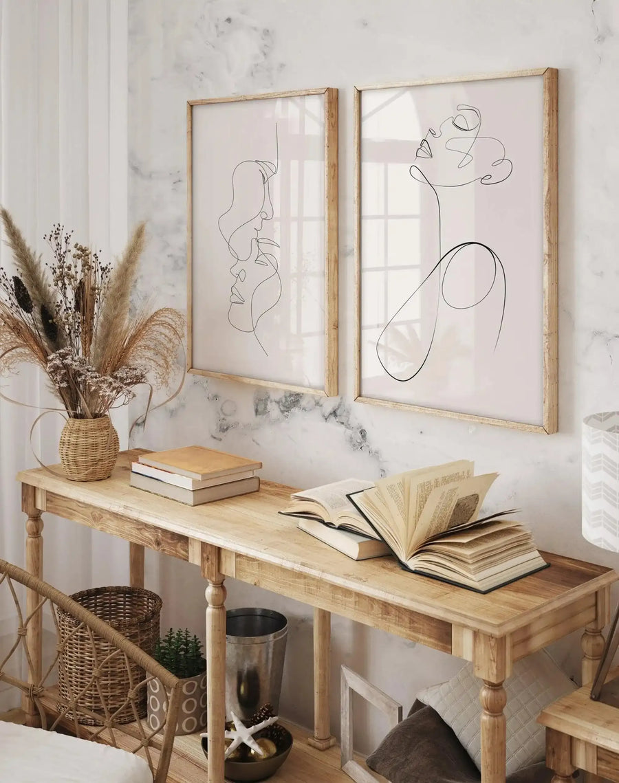 Two framed line art prints displayed above a wooden console table in a neutral-toned bedroom. The left print features two abstract faces in profile, while the right print shows a single abstract face looking upwards, both with black lines on a pale pink background and natural wood frames with white borders. The table holds a woven vase with dried pampas grass, stacked books, and an open book, with a woven basket and potted plant underneath.
