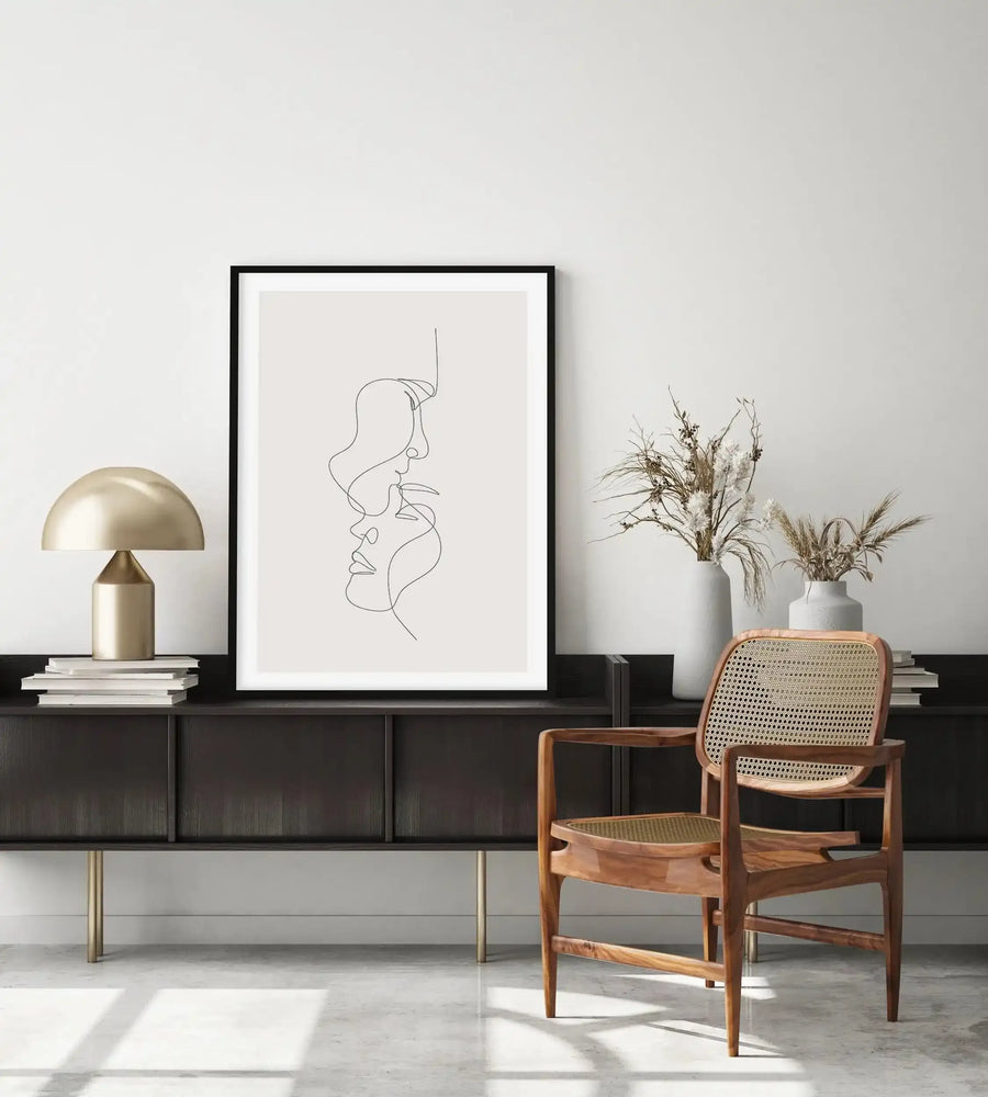 Lifestyle shot of a minimalist living room featuring a framed art print displayed above a dark wood console table. The print features a continuous line drawing of two faces in profile, one above the other, with soft curves and minimalist detail, in black on a light grey background, with a black frame and white border. The room includes a gold mushroom lamp, stacked books, dried floral arrangements in white vases, and a wooden chair with a woven cane back.