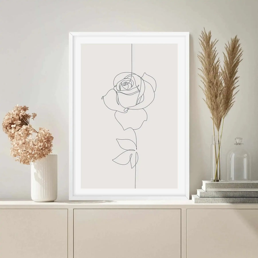 Framed art print displayed on a light cream console table in a minimalist room. The print features a single rose drawn in a continuous line art style, with a delicate black line forming the petals, stem, and two leaves against a pale grey background, with a white frame and white border. Dried hydrangeas in a white ribbed vase and pampas grass in a clear glass vase are also on the table.