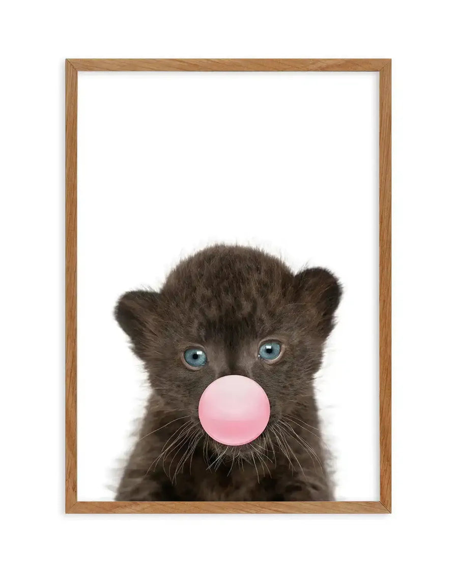 Art print in natural wood frame featuring a close-up portrait of a young black leopard cub with striking blue-grey eyes and detailed white whiskers against a white background. The cub blows a large, glossy bubble gum bubble in soft pink. The image has a playful, contemporary style with realistic fur texture and whimsical character.