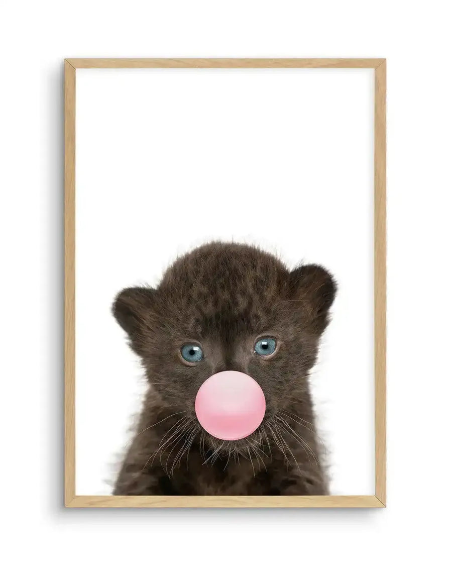 Art print featuring a close-up portrait of a young black leopard cub with striking blue-gray eyes against a white background. The cub has dark charcoal fur with visible texture, pointed ears, and delicate white whiskers. A glossy bubble gum pink sphere covers the cub's mouth, as if the animal is blowing a bubble. The photograph-style image is mounted in a natural wood frame with a white border.