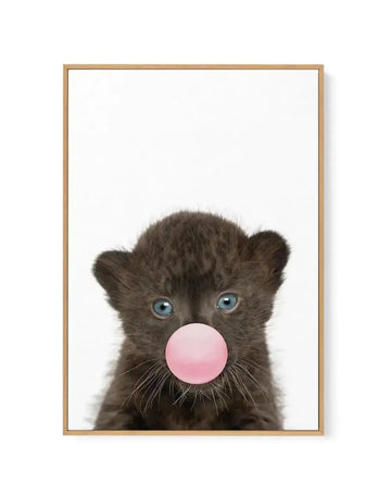 Framed canvas print featuring a close-up photograph of a black leopard cub with bright blue eyes, blowing a large pink bubble gum bubble, set against a stark white background. The cub's dark fur and delicate whiskers are clearly visible, with a natural wood frame.