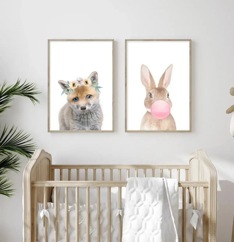 Framed art print displayed above a natural wood crib in a nursery setting. The print features a watercolor illustration of a young rabbit with tan and cream fur and pink inner ears, blowing a large bubble gum bubble in soft pink. The artwork is set against a white background with a natural wood frame and white border. A matching framed print of a young fox with golden fur wearing a delicate flower crown is displayed to the left. Green potted plants flank both sides of the wall display.