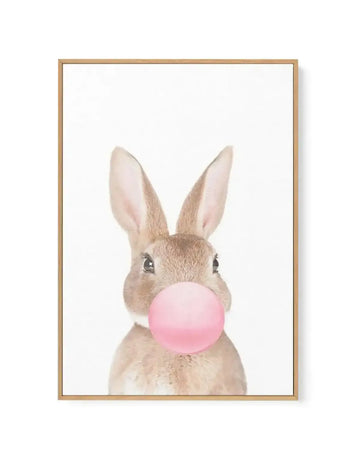 Framed canvas art print featuring an illustration of a young rabbit with tan and cream fur and long upright ears, blowing a large bubblegum pink bubble. The rabbit's dark eyes gaze forward against a clean white background. The artwork is presented in a natural wood frame with a white border.
