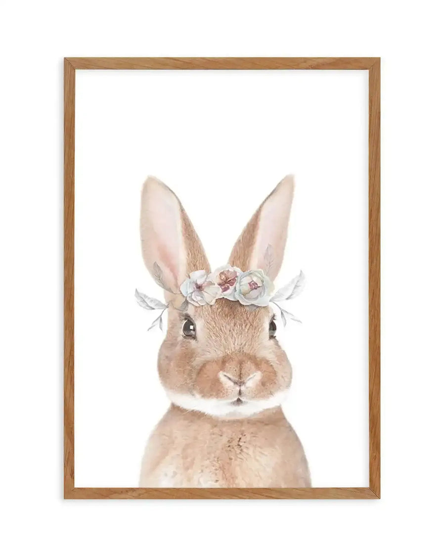 Watercolour art print featuring a fluffy brown bunny with large ears, wearing a delicate flower crown with soft pink, blue, and cream blossoms and green leaves, printed on fine art paper and displayed in a natural oak frame with white border.