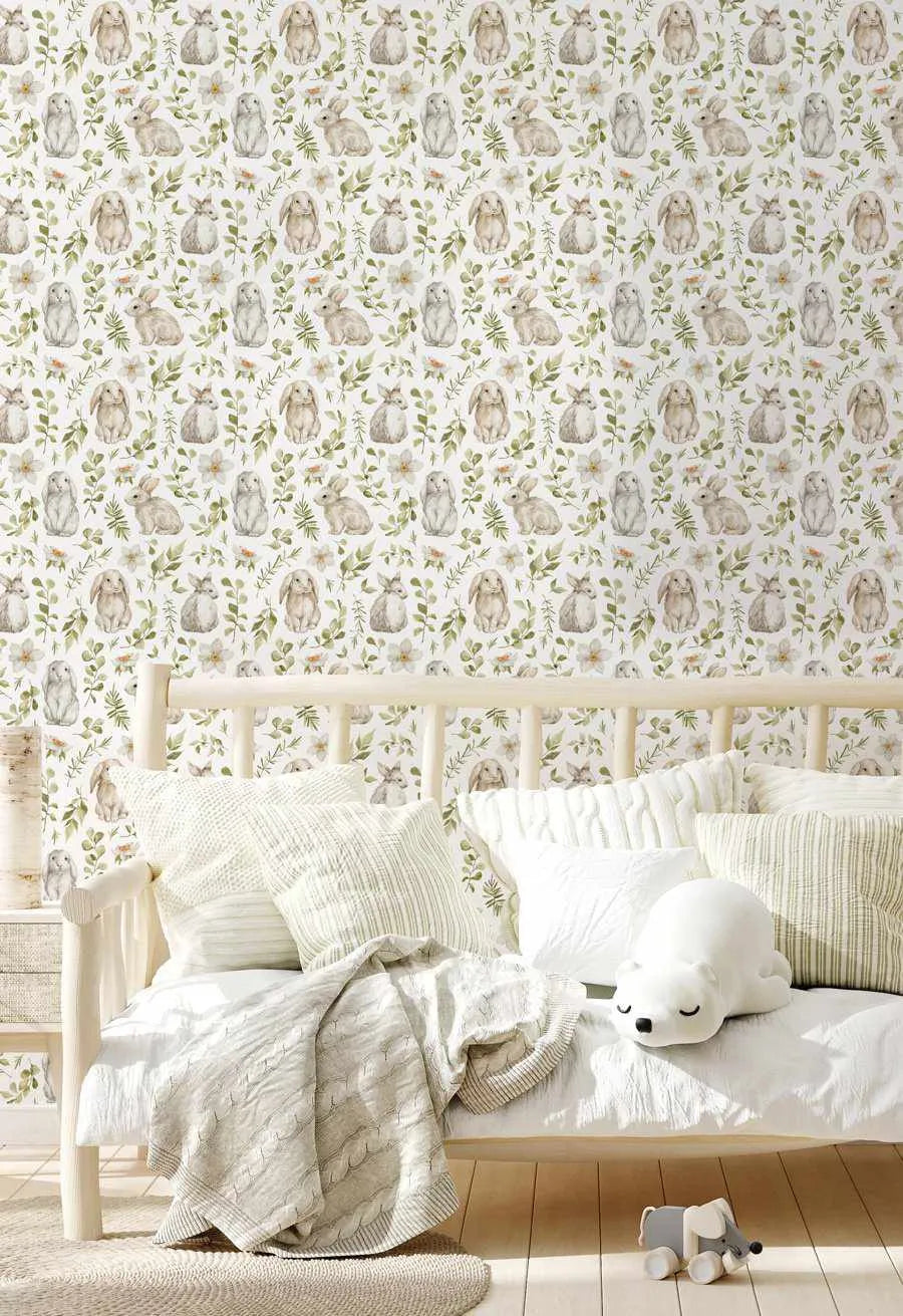 Lifestyle shot of a child's bedroom featuring Little Bunny wallpaper with a repeating pattern of hand-painted bunnies in various shades of cream, beige, and light brown, interspersed with green leafy branches and small white and pale yellow flowers on a white background. The wallpaper is displayed behind a light wood daybed with white bedding, several white and striped pillows, and a cable-knit throw blanket. A white polar bear plush toy and a wooden elephant toy are visible on the floor.