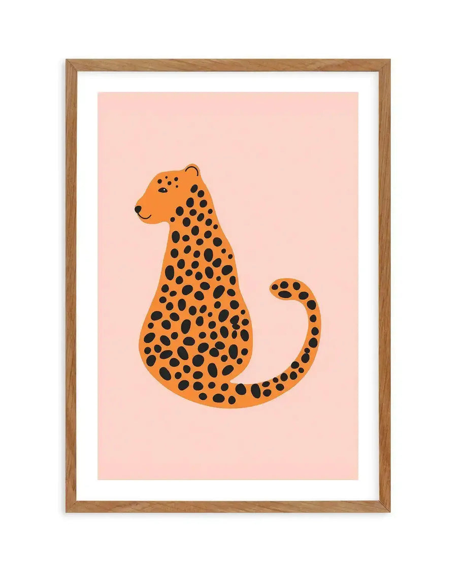 Art print featuring a stylized illustration of a cheetah sitting, facing left, with a long curved tail. The cheetah has an orange body with irregular black spots and a small smile, set against a soft blush pink background. The print is displayed in a natural oak frame with a white border.