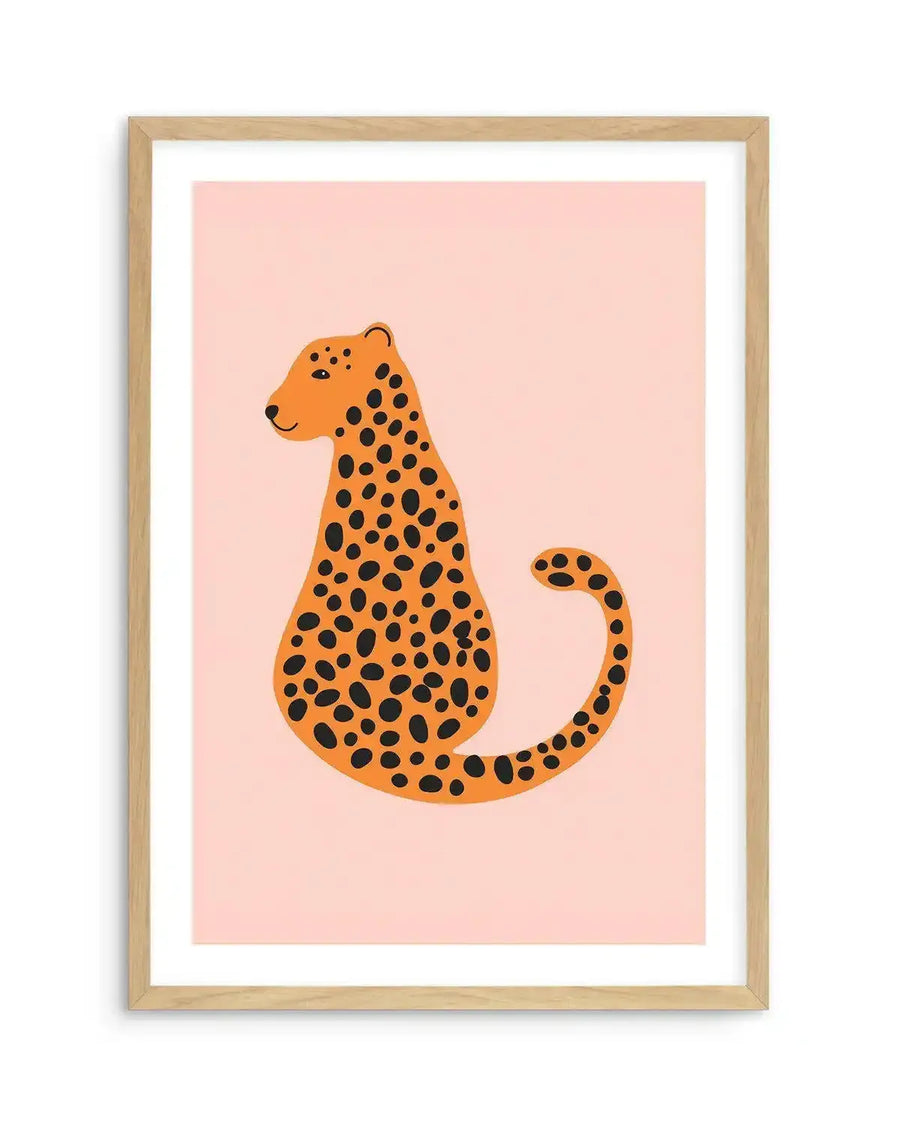 Art print featuring a stylized illustration of a sitting cheetah with a playful expression, rendered in warm orange with black spots on a soft blush pink background, displayed in a natural oak frame with white border.
