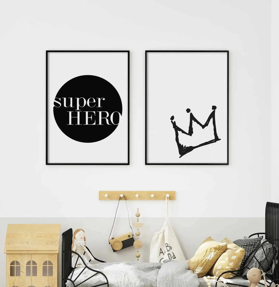 Two framed art prints displayed side-by-side above a child's bed in a minimalist bedroom. The left print features the words "super HERO" in white serif font on a solid black circle against a white background, with a black frame. The right print features a hand-drawn, brushstroke-style black crown illustration against a white background, with a black frame. The room includes a light wood peg rail with hanging items, a light wood dollhouse, and a black metal bed frame with white and yellow bedding.