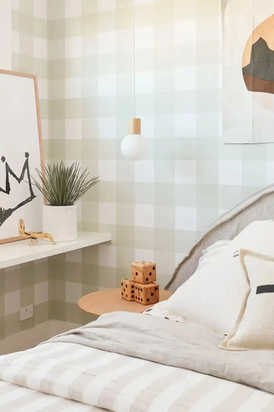 Framed art print featuring a black, hand-drawn crown with a slightly distressed, sketchy texture on a white background, displayed on a white shelf in a child's bedroom. The print has a natural wood frame and white border. The room features light green and white gingham wallpaper, a light wood bedside table with three stacked wooden dice, a white bed with striped bedding, and a small potted plant.