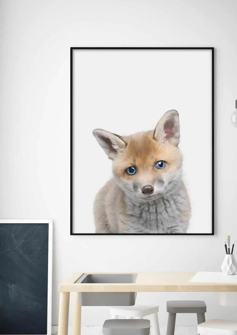Kids Room:
Framed art print of a baby fox displayed above a light wood children's desk in a bright, minimalist room. The print features a close-up of a young fox with soft brown fur, large pointed ears, and striking blue eyes, looking directly at the viewer with a slight head tilt. The print has a black frame and white border. The room includes a tall, dark grey chalkboard leaning against the wall and a white geometric vase with paintbrushes on the desk.