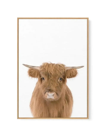 Framed canvas art print of a Highland cattle head against a white background. The animal features long, shaggy tan and brown fur, small curved horns extending outward, and a calm direct gaze. A white muzzle and gentle expression are visible. The painting has a soft, realistic style with warm earth tones. The canvas is displayed in a natural wood frame with a simple border.