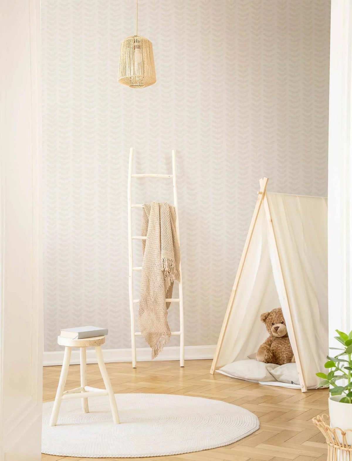 Little Leaf in Sand Wallpaper displayed on walls of a minimalist nursery room. The wallpaper features a subtle chevron or zigzag pattern in soft greige and cream tones with a matte finish. The room includes natural wood flooring, a white wooden ladder with a draped beige throw, a white stool with stacked books, a cream canvas teepee tent with a teddy bear inside, a woven pendant light, and potted greenery. The overall aesthetic is calm and neutral with warm, earthy tones throughout.
