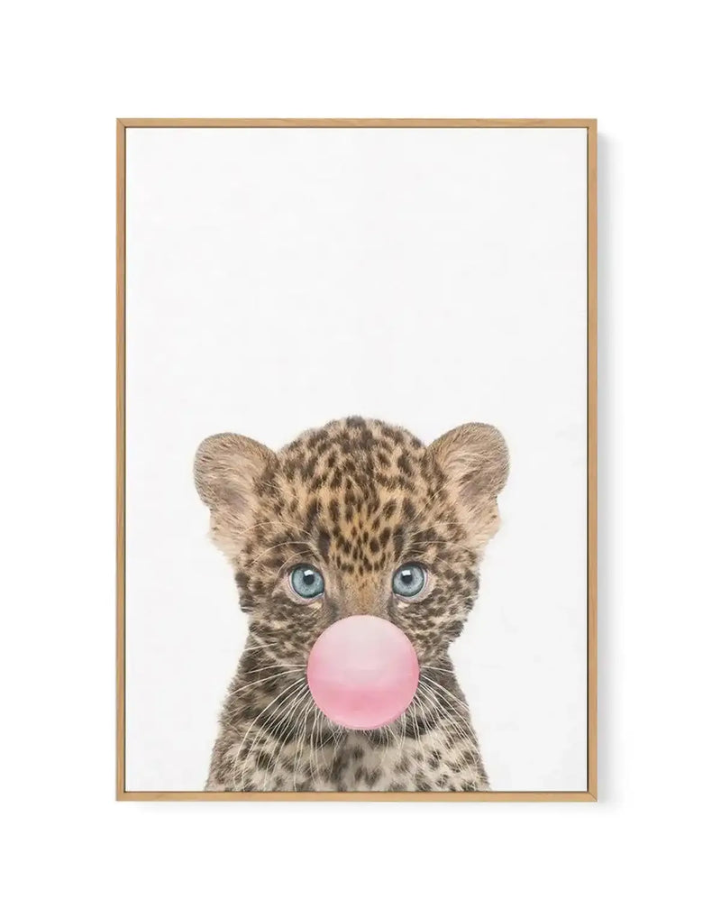 Framed canvas art print featuring a young leopard cub with spotted tan and brown fur and bright blue eyes, blowing a large bubble gum pink bubble. The cub is centered against a clean white background in a portrait orientation. The artwork has a soft, illustrated style with detailed fur texture. The canvas is mounted in a natural wood frame with a thin border, displayed against a white wall with subtle shadow visible on the right edge.